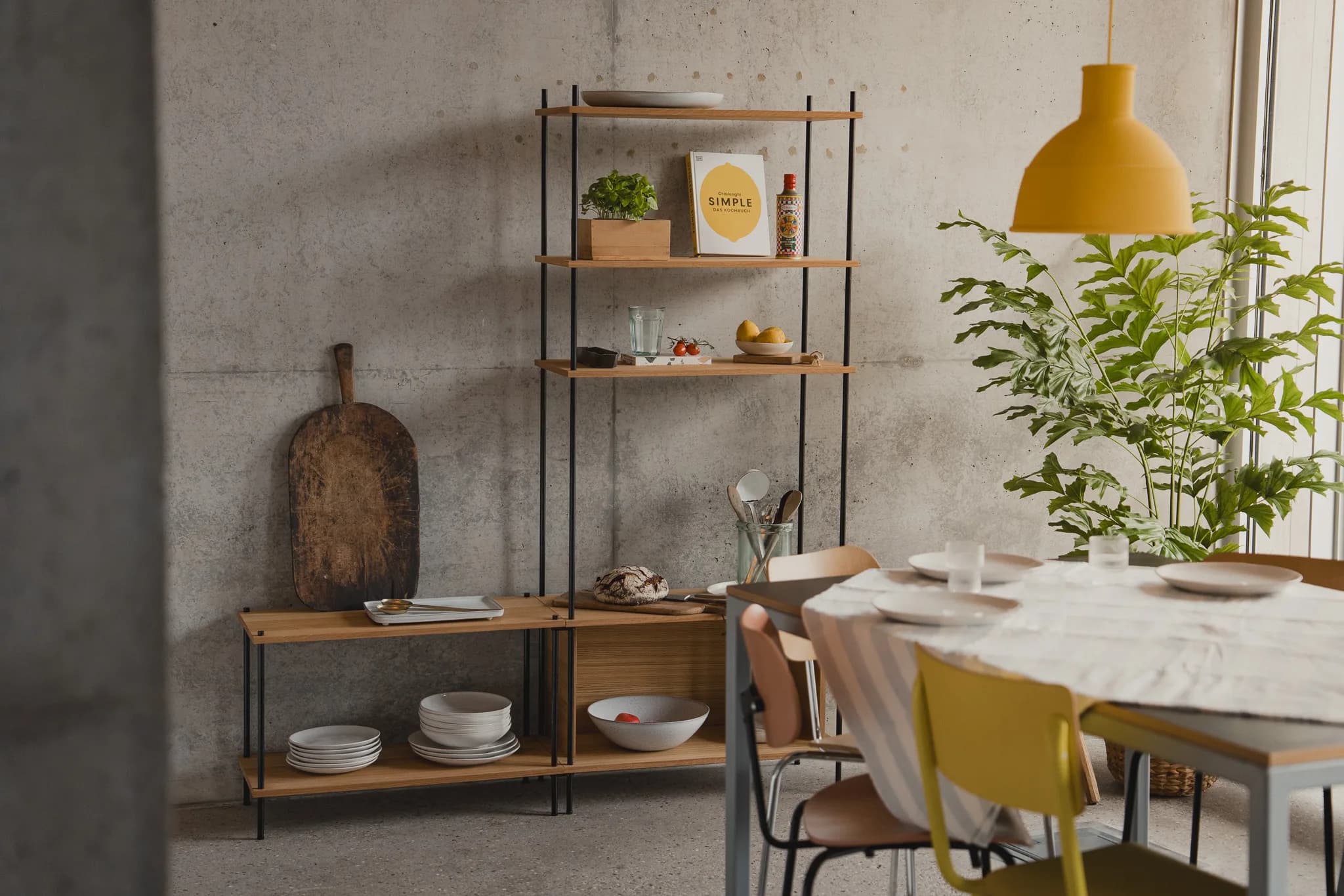 Loft-Esszimmer mit hohem KUUDU-Standing-Regal voller Kochbücher und Vorrat, davor gedeckter Esstisch mit gelber Pendelleuchte und großer Pflanze